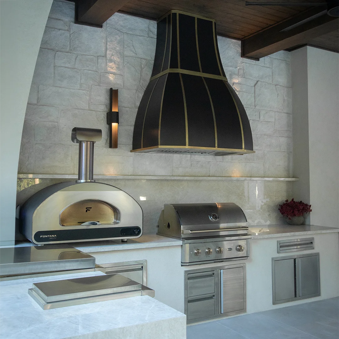 Outdoor kitchen setup with a black and gold range hood, a Fontana Volta 90 stainless steel pizza oven, a stainless steel Wildfire gas grill, and built-in cabinets against a light stone tile wall.