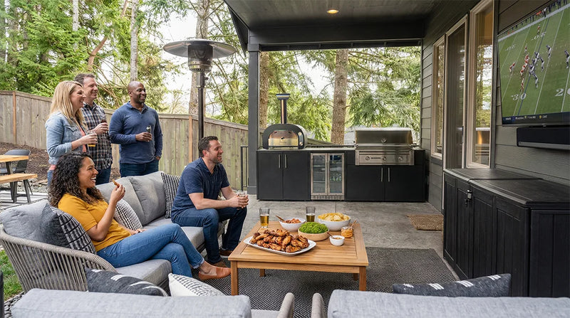 Group of people socializing outdoors in a backyard with the Elysia outdoor kitchen, Mastro 60 oven, Grill, TV, and patio furniture.